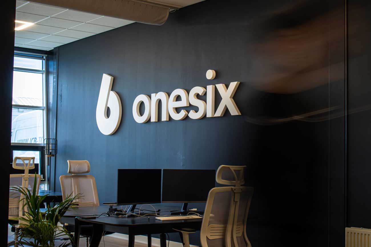 Office interior with a black wall displaying a large white 'onesix' logo above a desk with two monitors and ergonomic chairs.
